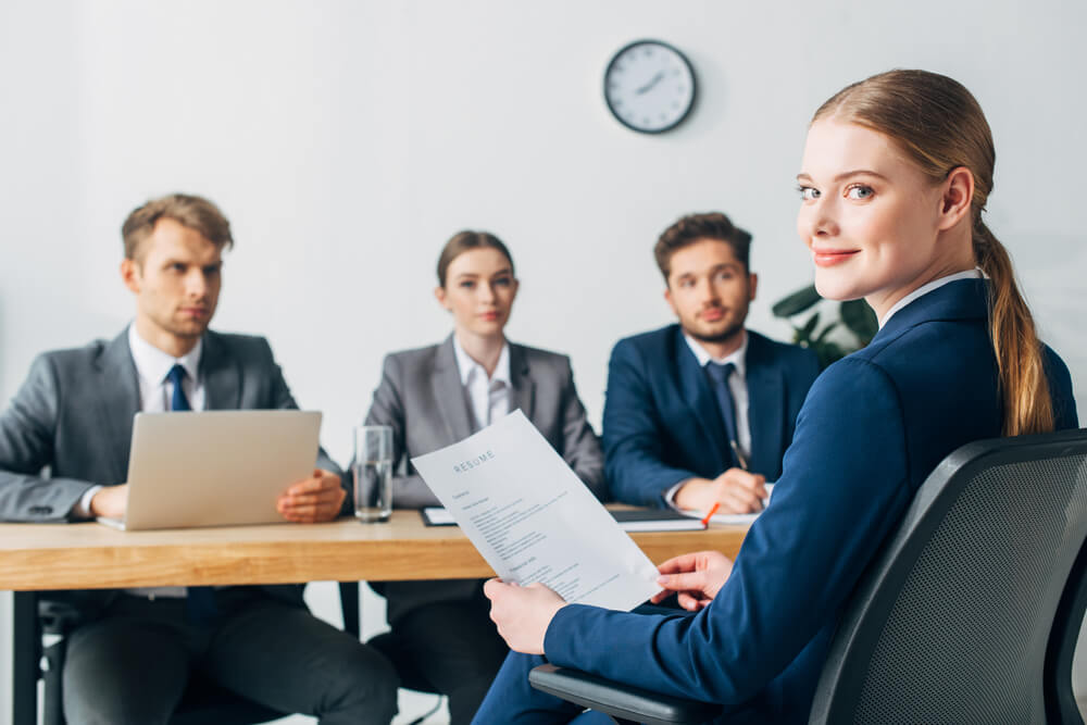 female job applicant looking at camera while holding resume near three recruiters